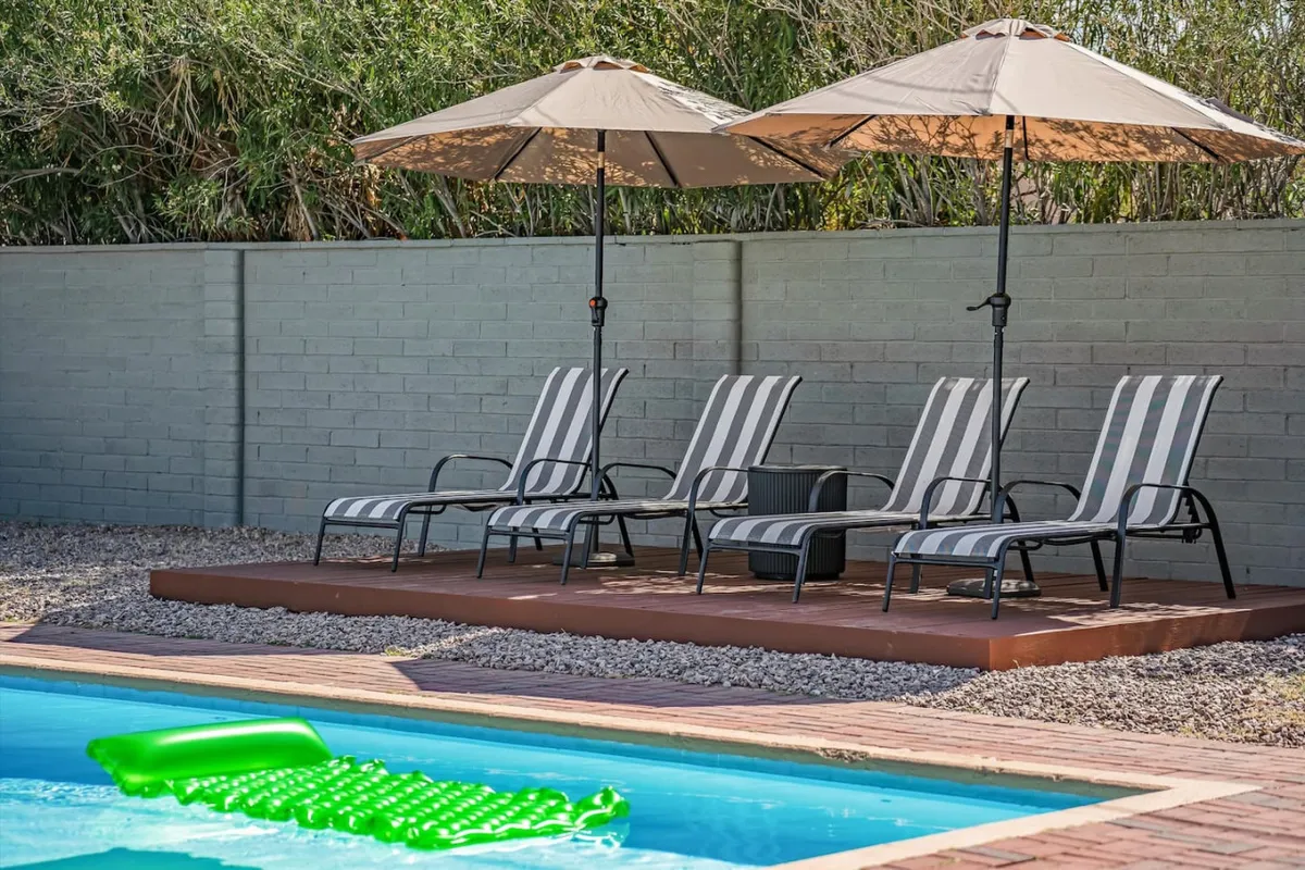 This image features a sparkling swimming pool flanked by lounge chairs and beach umbrellas, with a green pool float adding a playful touch. The surrounding area is bright and inviting, suggesting a space ideal for both active play and leisurely relaxation. The landscaping hints at a Southwest vibe, enhancing the desert retreat ambiance.