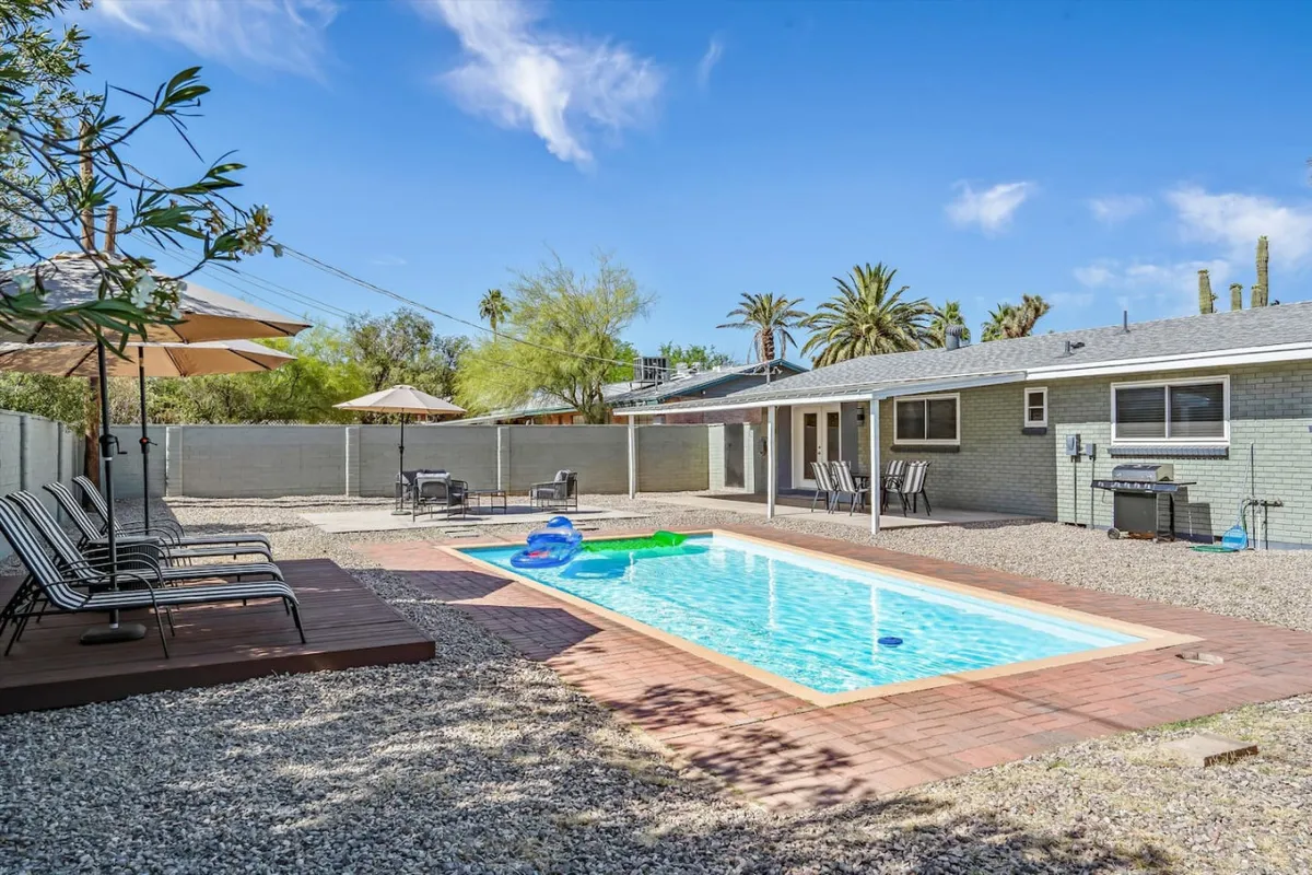 The photo focuses on an inviting swimming pool bordered by outdoor seating, palm trees, and a large umbrella. The setup emphasizes both sunbathing and shaded relaxation, with space for gatherings and fun activities, set against a backdrop of desert plants.