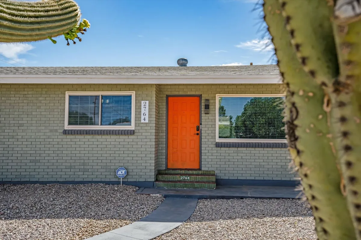 A vibrant orange front door framed by a cactus plant stands out against the gray brick exterior, creating an inviting entrance. The scene suggests a welcoming atmosphere for guests arriving to start their Tucson adventure.