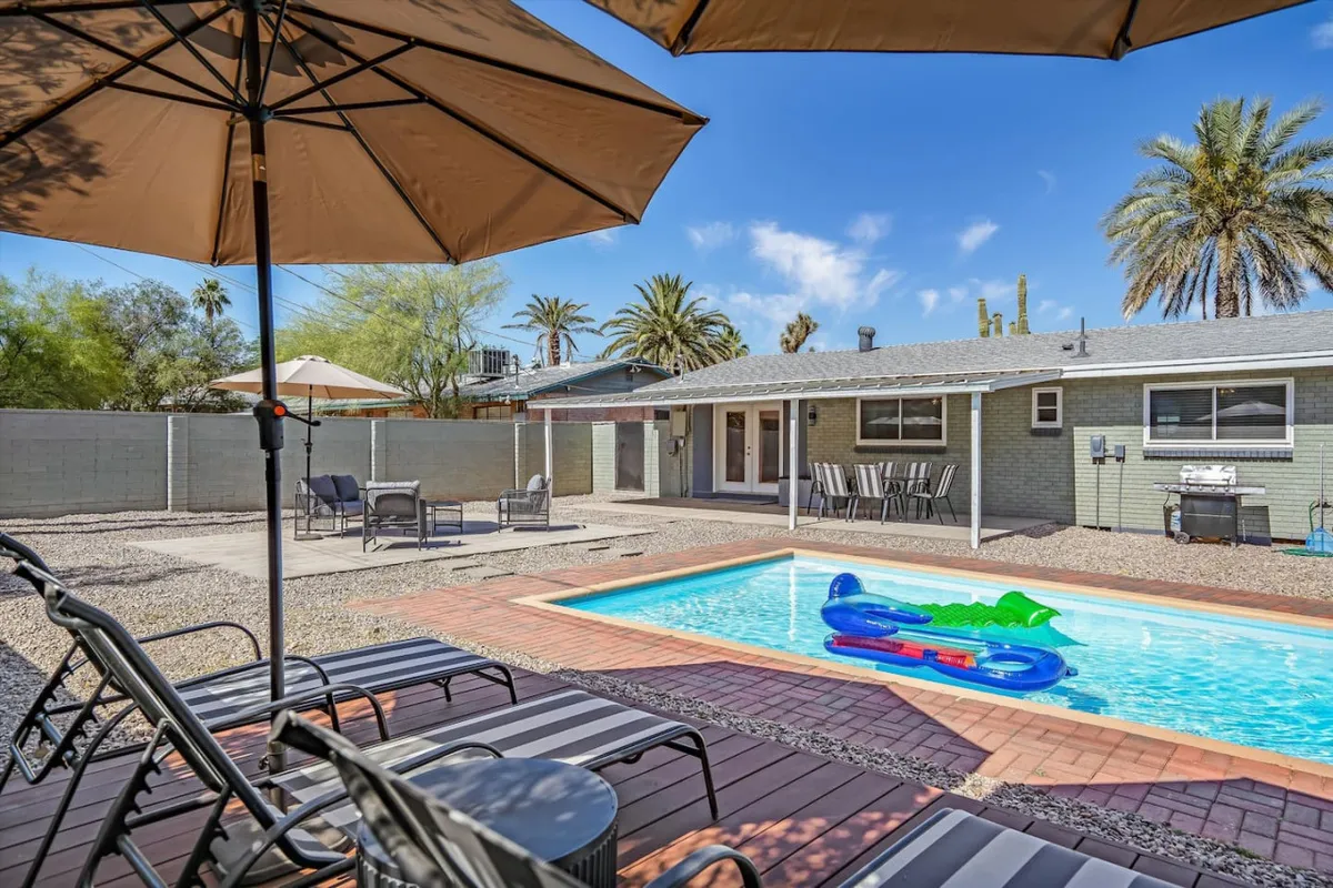The hero image showcases the sunlit backyard of a single-family home in Tucson, Arizona, featuring a sparkling swimming pool surrounded by inviting lounge chairs and umbrellas, a shaded outdoor dining area, swaying palm trees, and a built-in backyard grill. The scene radiates relaxation and fun, with hints of sunset hues and ample space for families or groups to gather and unwind.