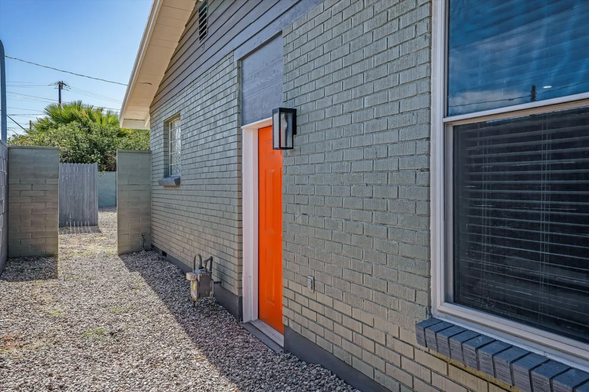 A bright and cheerful exterior scene with a textured green wall, a striking orange door, and a window with blinds. The focus is on the entrance, echoing the sense of community and the welcoming nature of the property.