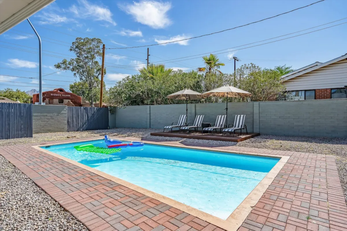 A sunny outdoor pool area featuring inviting lounge chairs, umbrellas for shade, and lush landscaping. The scene is bright and leisurely, with clear blue water and plenty of space for relaxing or gathering after a day outdoors.