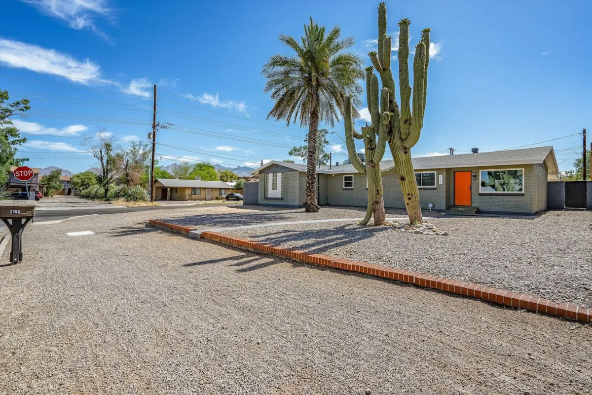 The scene shows a cactus and palm tree in a gravel driveway, with an orange door in the background. The space looks perfect for group gatherings or outdoor relaxation, with a distinct Southwest landscape that matches the described desert hardscape. The inviting mood and spacious yard suggest opportunities for kids and pets to play freely.