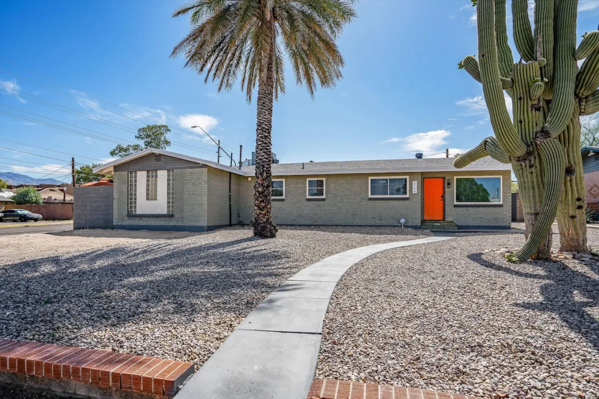 This vibrant exterior shot showcases a bright orange door framed by a tall palm tree, a large cactus, and a well-maintained gravel landscape. The desert rock hardscape is prominent, providing a clear view of the secure and pet-friendly outdoor areas.