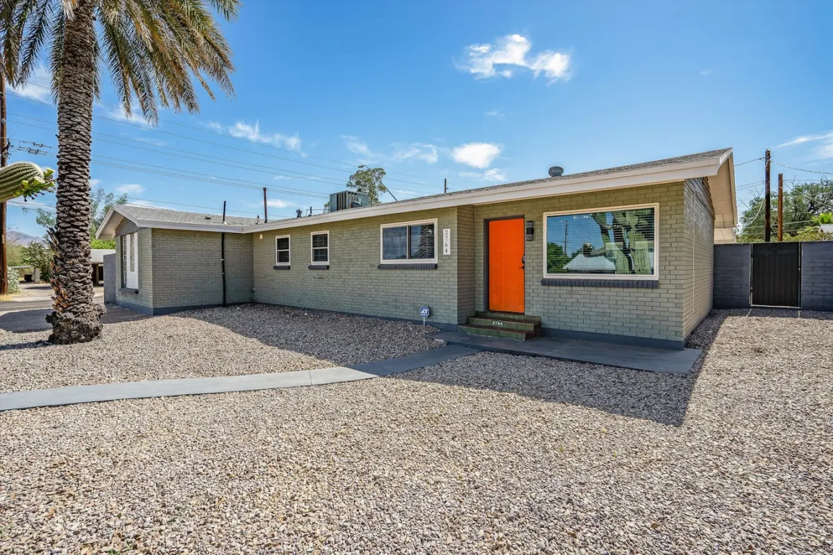 The orange front door is set against a landscaped gravel yard with palm trees, creating a tranquil and distinctly Southwestern outdoor setting ideal for relaxing or socializing outdoors.