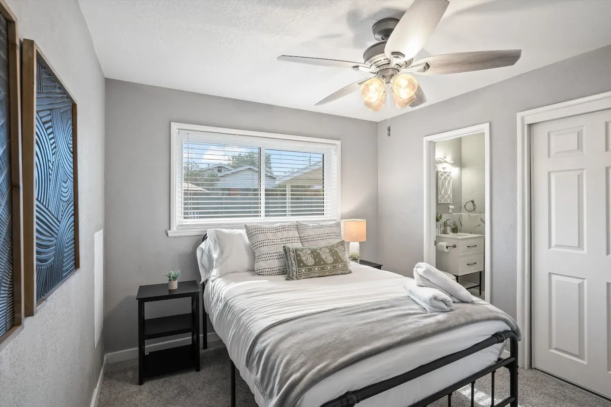 A tranquil bedroom with a bed adorned with decorative pillows, bathed in natural light from a nearby window. A nightstand with a lamp sits beside the bed, enhancing the cozy and serene atmosphere. The room feels perfect for relaxing and unwinding.