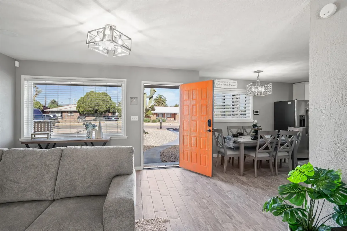 This image captures the open-concept heart of the home with a bright orange door, a welcoming dining table, a comfortable sofa, and abundant natural light streaming through the windows. The arrangement visually connects the living and dining spaces, supporting family gatherings and social interaction.