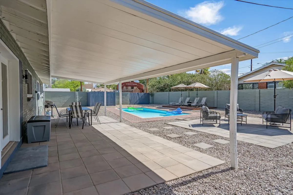 This image captures a sparkling swimming pool surrounded by lounge chairs, with a covered patio and an outdoor dining set nearby. The area is enclosed, features desert-style landscaping with rocks, and is bathed in sunlight, evoking a leisurely and carefree mood ideal for both family play and adult relaxation.